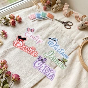 May include: Five embroidered name patches featuring Disney-inspired motifs. The patches display the names Emily, Daniel, William, James, and Olivia in various colours and fonts, accented with icons like Mickey ears, bows, and hats on a fabric surface.