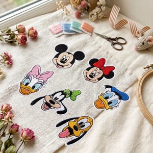 May include: A collection of six embroidered character patches featuring Mickey Mouse, Minnie Mouse, Daisy Duck, Goofy, Donald Duck, and Pluto arranged on a piece of light beige fabric, accompanied by embroidery thread spools and scissors.