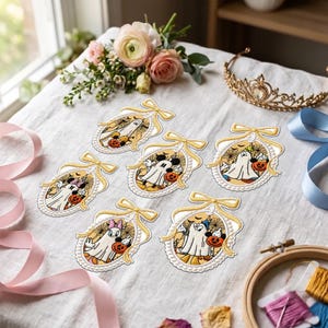 May include: Six oval-shaped embroidered patches featuring cartoon characters dressed as ghosts for Halloween. Each patch is framed in gold thread with a bow at the top and pearl-like accents, depicting characters holding orange jack-o'-lanterns.