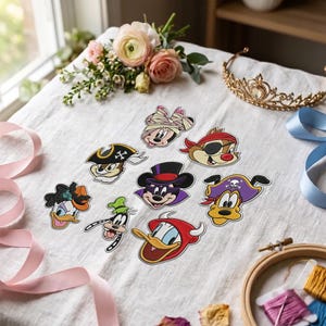 May include: A collection of eight embroidered patches featuring cartoon character heads in various costumes, including a mummy, a pirate, a magician, and a devil, arranged on a white fabric surface with flowers, a tiara, and ribbons.