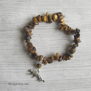 Könnte beinhalten: Armband aus polierten Tigerauge-Steinen in Braun- und Goldtönen. Ein silberfarbener Tiger-Anhänger und eine kleine Perle hängen am Armband. Die Website-Adresse blaqkat.etsy.com ist sichtbar.