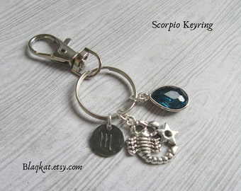 Puede incluir: Llavero plateado con un mosquetón, un anillo y tres colgantes. Los colgantes incluyen una gema en forma de lágrima de color azul oscuro, un disco redondo con el símbolo de Escorpio y un escorpión con una estrella. El texto "Scorpio Keyring" es visible.