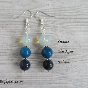 Könnte beinhalten: Ein Paar silberne Ohrringe mit jeweils drei Perlen. Die obere Perle ist opaleszierend weiß, die mittlere Perle ist aus blauem Achat und die untere Perle ist aus Sodalith.