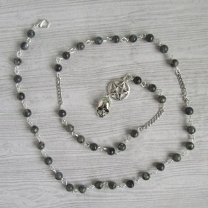 Puede incluir: Un collar de cuentas negras con un dije de plata en forma de calavera y pentagrama. El collar está hecho de cuentas negras y cadena de plata.
