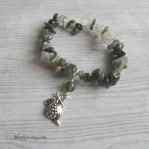 Könnte beinhalten: Ein Armband mit Edelsteinen in Grün, Grau und Klar. Ein silberner Anhänger in Form einer Frucht hängt am Armband. Das Armband wird auf einer hellen Holzmaserung präsentiert.