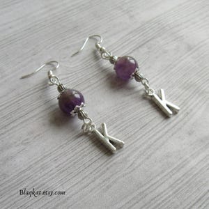 Puede incluir: Pendientes colgantes con una cuenta de amatista púrpura y un dije de letra "K" plateada. Los pendientes tienen ganchos plateados y detalles metálicos decorativos. Los pendientes se exhiben sobre una superficie de madera gris claro. El nombre de la tienda Etsy "Blaqkat.etsy.com" es visible.