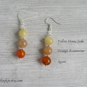 Könnte beinhalten: Ein Paar silberne Ohrringe mit jeweils drei Perlen. Die obere Perle ist gelb, die mittlere Perle ist orange und die untere Perle ist braun. Die Perlen sind mit "Gelber Honig-Jade", "Orange Aventurin" und "Achat" beschriftet.