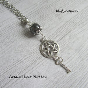 May include: A silver chain necklace with a black gemstone pendant, a silver pentagram charm, and a silver key charm. The necklace is on a light wood background. The text "Goddess Hecate Necklace" is below the necklace.  The website address "Blagkat.etsy.com" is in the top right corner of the image.