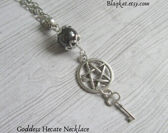Puede incluir: Un collar de cadena de plata con un colgante de piedra preciosa negra, un amuleto de pentagrama de plata y un amuleto de llave de plata. El collar está sobre un fondo de madera clara. El texto "Goddess Hecate Necklace" está debajo del collar. La dirección del sitio web "Blagkat.etsy.com" está en la esquina superior derecha de la imagen.
