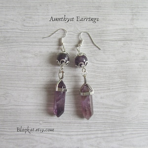 Könnte beinhalten: Amethyst-Ohrringe mit silberfarbenen Metallakzenten. Jeder Ohrring hat einen facettierten Amethystkristallanhänger und eine runde Amethystperle. Die Ohrringe hängen an silberfarbenen Ohrhaken. Der Text "Amethyst Earrings" und "Blaqkat.etsy.com" sind sichtbar.