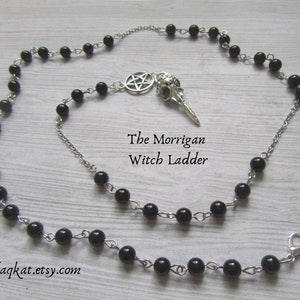 Puede incluir: Un collar de escalera de bruja plateado con cuentas negras. El collar presenta un colgante de calavera de cuervo y un dije de pentagrama. El texto "The Morrigan Witch Ladder" y "blaqkat.etsy.com" son visibles.