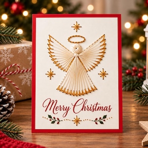 May include: A rectangular greeting card with a red border on a white background featuring a string art angel design in cream and gold thread. The text Merry Christmas is printed in red script below the angel, flanked by small green leaf accents.