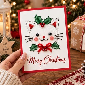 Puede incluir: Una tarjeta de felicitación blanca con borde rojo que presenta un diseño de arte con hilos de una cara de gato. El gato tiene ojos de cuentas negras, nariz roja, mejillas bordadas en rosa y bigotes negros. Está decorado con hojas de acebo y un lazo rojo.