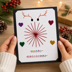 Puede incluir: Una tarjeta de felicitación rectangular con borde negro sobre fondo blanco. Presenta un patrón de rayos de sol bordado con hilo rojo, dos puntos negros para los ojos, astas bordadas en naranja, cuatro corazones brillantes y el texto MERRY CHRISTMAS en cuentas.
