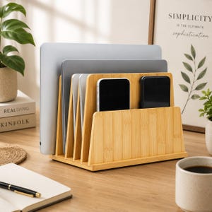 May include: A bamboo desktop organiser featuring five vertical slots of varying heights. The organiser holds a large silver laptop, a smaller silver tablet, and two black smartphones upright. The natural wood grain is visible throughout the structure.