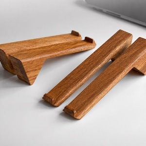 Può includere: Due paia di supporti inclinati per computer portatile in legno con una finitura naturale a venature marroni. Ogni supporto presenta una base a forma di cuneo e un bordo superiore dentellato per sostenere un computer portatile.