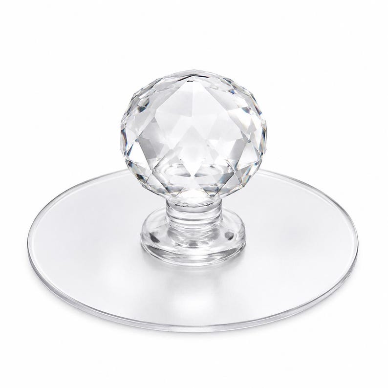 May include: A clear, faceted crystal-style spherical knob attached to a circular, transparent flat base. The knob features a multi-faceted geometric surface that reflects light, while the base is smooth and round, providing a stable foundation.