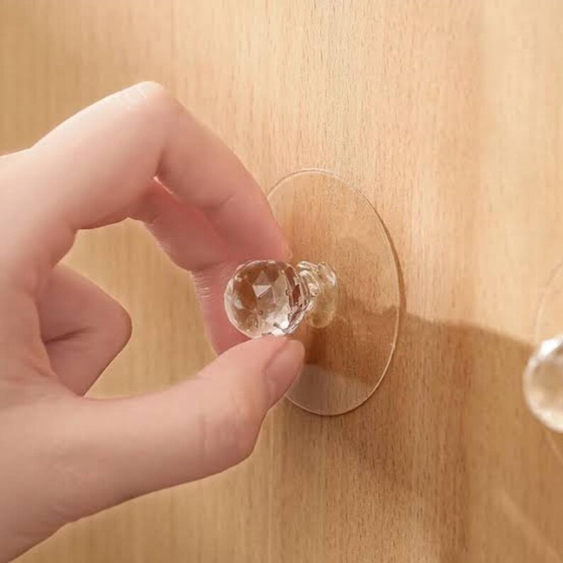 May include: A clear, faceted, crystal-like knob attached to a circular, transparent adhesive backing. The knob features a multi-sided geometric shape that reflects light, designed for mounting onto a flat surface for use as a handle or hook.