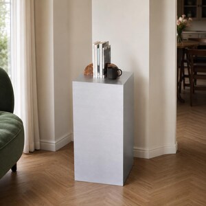 May include: A tall, rectangular silver pedestal stand with a smooth, matte finish. On top of the pedestal sits a curved wooden bookend, a small collection of upright books with white and black spines, and a solid black ceramic coffee mug.