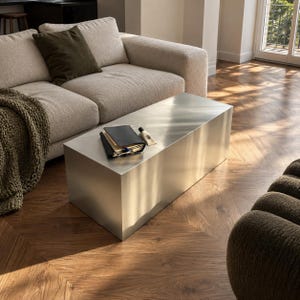 May include: A rectangular coffee table with a smooth, brushed silver metal finish. The table has a clean, boxy silhouette with sharp edges and a flat top surface. A black notebook and a small tube are placed on the center of the table surface.