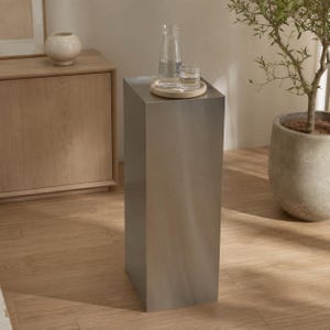 Könnte beinhalten: Ein hoher, rechteckiger Sockel mit einer Oberfläche aus gebürstetem silberfarbenem Metall. Auf der flachen Oberseite steht ein rundes Holztablett mit einer klaren Glaskaraffe mit Wasser und einem passenden Glasbecher in schlichter, zylindrischer Form.