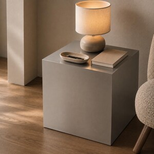 May include: A metallic silver cube side table with a smooth finish. On top sits a beige table lamp with a cylindrical shade, a small oval-shaped beige tray containing a black pen, and a rectangular beige book with a plain cover.