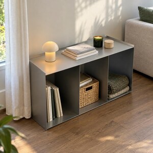 May include: A rectangular gray metal storage unit with three open compartments. The top surface holds a small mushroom-shaped lamp, a stack of books, a dark green candle, and a small round container. Inside are books, a woven basket, and folded textiles.