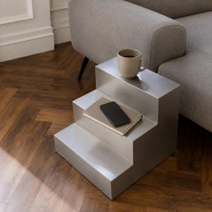May include: A metallic silver step-shaped side table with a smooth finish. A beige ceramic mug filled with a dark liquid sits on the top tier, while a closed book with a black smartphone resting on its cover is placed on the middle tier.