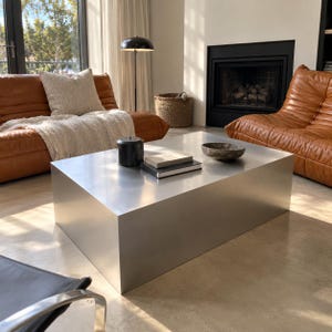 May include: A rectangular coffee table with a smooth, brushed silver metal finish. The table surface holds a small black cylindrical container, two stacked books with white pages, and a shallow, dark-toned circular bowl made of stone.