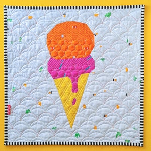 Op de afbeelding: Een wit gestikt vierkant van stof met een zwart-witte rand. Een kleurrijk ijshoorntje is in het midden aangebracht. Het ijshoorntje is gemaakt van oranje, roze en gele stof.