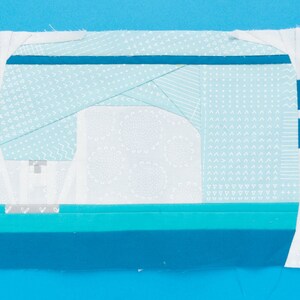 May include: A white fabric square with a blue and white patterned design. The fabric is sewn together in a patchwork style, creating a geometric design. The fabric is folded in half and the edges are sewn together.