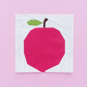 Op de afbeelding: Een rode appel met een groen blad en een bruine steel, gemaakt van stofresten, op een roze achtergrond.