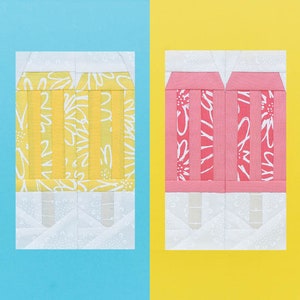 Pode incluir: Dois blocos de tecido em forma de picolé, um amarelo com estampa floral branca e outro rosa com estampa floral branca. Os picolés são feitos de quadrados de tecido costurados juntos para criar um visual acolchoado.