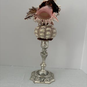 Pode incluir: Um objeto decorativo com uma base de castiçal de metal prateado com um pé hexagonal ornamentado. Sobre o castiçal repousa um ouriço-do-mar redondo e texturizado, encimado por uma grande concha espinhosa com um interior cor-de-rosa.
