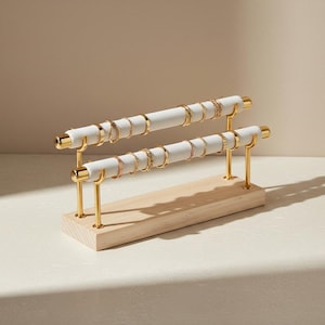 May include: A two-tier jewellery display stand featuring two horizontal white fabric-covered bars supported by gold-toned metal posts on a rectangular light-coloured wooden base. Several gold rings are arranged along the two padded bars.