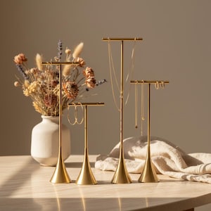 May include: Four gold-toned metal jewelry stands of varying heights with T-shaped bars. The stands feature slender stems and conical bases, displaying gold necklaces and hoop earrings. The metal finish is smooth and reflective, providing a structured display for various pieces of jewelry.
