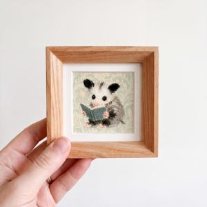 May include: A small square wooden frame containing an embroidered artwork of a white and grey opossum reading a teal book. The opossum has pink paws and ears, and the background features a light green floral pattern on a cream-coloured fabric.