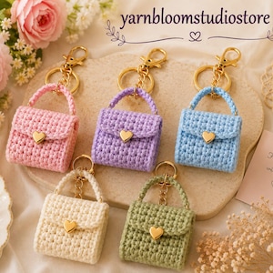 Pode incluir: Cinco bolsas de crochê em miniatura nas cores rosa, roxo, azul claro, creme e verde sálvia. Cada bolsa possui um fecho dourado em formato de coração e um chaveiro de metal dourado. O texto yarnbloomstudiostore e Handmade with love é visível.