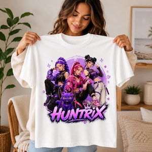 Può includere: Una t-shirt bianca a maniche corte con una stampa grafica di tre personaggi in stile anime e un gatto viola. Il design include il testo "HUNTRIX" in grassetto viola, circondato da stelle e cuori su uno sfondo circolare di colore viola.