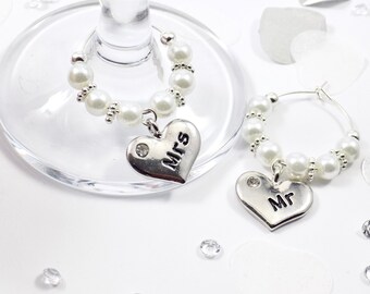 Könnte beinhalten: Zwei silberne herzförmige Weinglas-Anhänger mit den Worten "Mr." und "Mrs." darauf. Jeder Anhänger ist an einem silbernen Ring mit weißen Perlen befestigt.