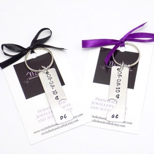 May include: Two personalized keychains with stamped dates and initials, attached to keyrings. Each keychain is paired with a card and tied with a black or purple ribbon. The keychains have the date 06.08.10 and initials.