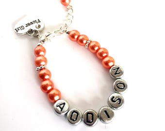 Könnte beinhalten: Ein Armband mit orangefarbenen runden Perlen und silberfarbenen Buchstabenperlen, die den Namen ADDISON bilden. Das Armband enthält einen silberfarbenen herzförmigen Anhänger mit der Gravur Flower Girl und eine silberfarbene Metallkettenverlängerung.