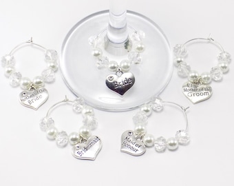 May include: Five silver-toned metal wine glass charms featuring white faux pearls and clear faceted beads. Each charm has a heart-shaped pendant engraved with the words Bride, Mother of the Bride, Mother of the Groom, Bridesmaid, or Maid of Honour.
