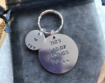 May include: A silver-toned metal keychain featuring two circular charms attached to a split ring. The larger charm is engraved with the text THIS DADDY BELONGS TO... and the smaller charm is engraved with a heart symbol above the name IVY.