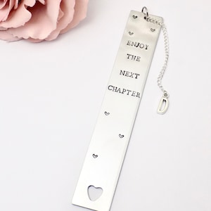 May include: Silver bookmark with the text "Enjoy the next chapter" and a heart cut-out at the bottom. A silver chain with a letter "D" charm is attached to the top.
