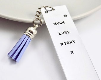 May include: A rectangular silver-toned metal bookmark featuring the stamped black text "MUCH LOVE NICKY x" and small star icons. A silver jump ring connects the bookmark to a light purple suede-style tassel with a polished silver-toned cap.