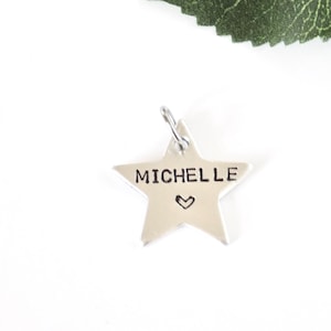 May include: A silver star-shaped pendant with the name "MICHELLE" stamped on it, and a small heart symbol. The pendant has a loop at the top for hanging. A green leaf is visible in the background.