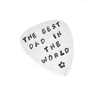 Puede incluir: Una púa de guitarra plateada con la frase "THE BEST DAD IN THE WORLD" en letras negras. Una pequeña estrella en la parte inferior. La púa está sobre un fondo blanco.