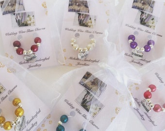 Könnte beinhalten: Fünf Hochzeits-Weinglasanhänger in weißen Organzabeuteln auf Karten mit der Aufschrift Wedding Wine Glass Charms. Die Anhänger bestehen aus farbigen Perlen und silberfarbenen herzförmigen Plaketten mit Texten wie Bride und Bridesmaid.