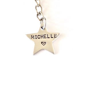 May include: A silver-colored star-shaped charm with the name "MICHELLE" stamped on it, along with a small heart symbol. The charm is attached to a silver chain, creating a personalized accessory.
